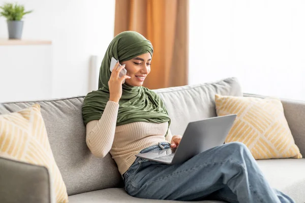 Joven mujer árabe alegre en hijab usando el ordenador portátil para el trabajo a distancia, llamando en el teléfono inteligente, comunicándose en línea desde casa — Foto de Stock