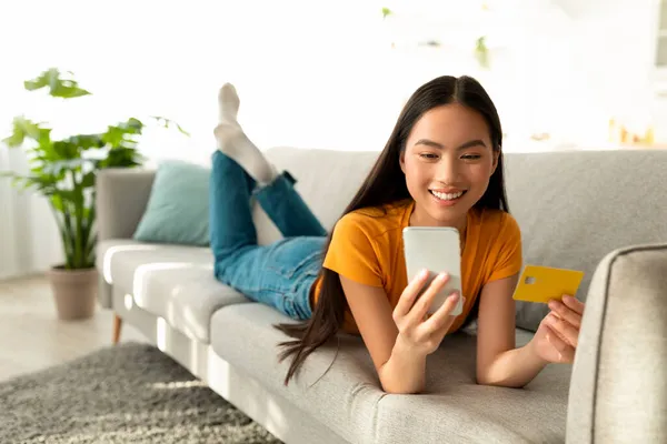 Pleasant feelings from buying. Happy asian woman looking at smartphone and using credit card, lying on sofa and smiling — Stock Photo, Image