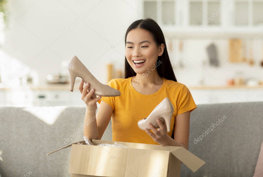 Happy asian woman received package, unpacking cardboard box, holding and looking at high heel shoes, sitting on sofa