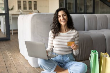 Mutlu genç bayan evde online alışveriş yapıyor, kredi kartı ve laptop kullanıyor, para transfer ediyor, internetten mal sipariş ediyor.