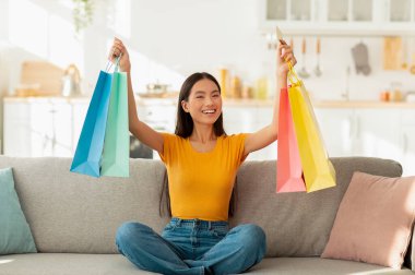 Tatmin olmuş Asyalı kadın renkli alışveriş çantaları ve kredi kartıyla kanepede oturuyor, evden online alışveriş yapıyor.