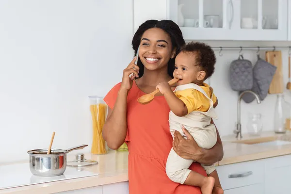 Alegre negro mujer hablando en celular en cocina con bebé en manos —  Fotos de Stock