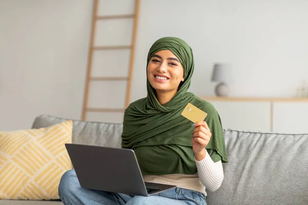 Güzel genç Arap kadın tesettürde oturuyor, kredi kartı ve laptop kullanıyor, kanepede oturuyor, internetten mal alıyor. — Stok fotoğraf