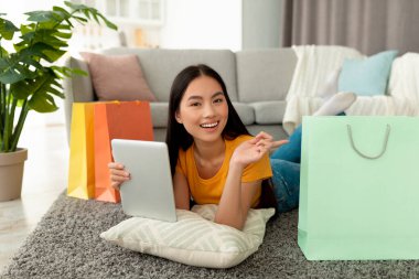 Alışverişte ve e-ticarette. Tablet tutan mutlu Asyalı kadın, internetten alışveriş yapıyor, yerde yatıyor ve hediye çantasına işaret ediyor.