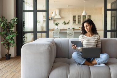 Genç beyaz kadın dizüstü bilgisayar ve dijital tablet kullanıyor, çalışıyor ya da çalışıyor, evdeki kanepede oturuyor, boşluğu kopyalıyor.
