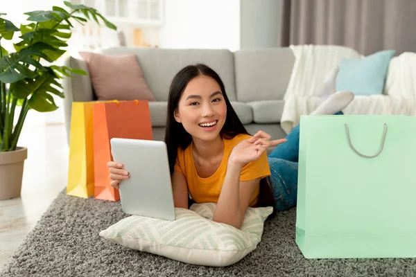 Shopaholism y e-commerce. Mujer asiática feliz sosteniendo la tableta, compras en línea, acostado en el suelo y apuntando a la bolsa de regalo — Foto de Stock