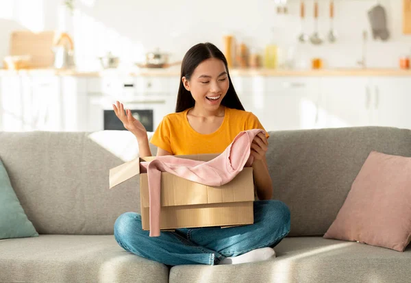 Objednávka oblečení online. Šťastný korejský zákazník otevírá balíček, dívá se na novou košili, sedí doma na pohovce — Stock fotografie