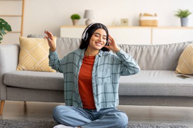 Olumlu genç bir Arap kadın evde oturmuş, kulaklık takıyor, müzik dinliyor ya da sesli kitap dinliyor.