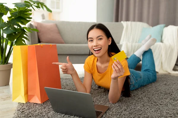 Emocionado mujer asiática comprar en línea con tarjeta de crédito y portátil, apuntando a las bolsas de la compra, acostado en el suelo en casa — Foto de Stock