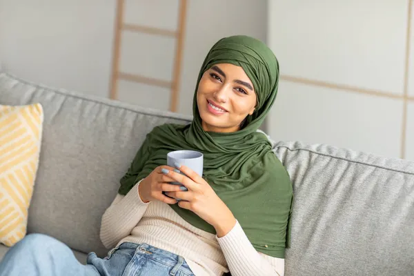 穿着头巾在家里沙发上喝咖啡的快乐阿拉伯妇女的画像 — 图库照片