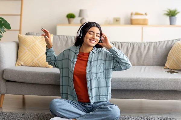 Θετική νεαρή Άραβα γυναίκα κάθεται στο πάτωμα με ακουστικά, ακούγοντας μουσική ή ακουστικό βιβλίο στο σπίτι — Φωτογραφία Αρχείου