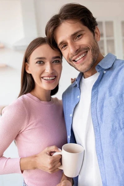 Portret wesołej, uśmiechniętej młodej żony i męża z filiżanką gorącego napoju robiących zdjęcia w kuchni — Zdjęcie stockowe