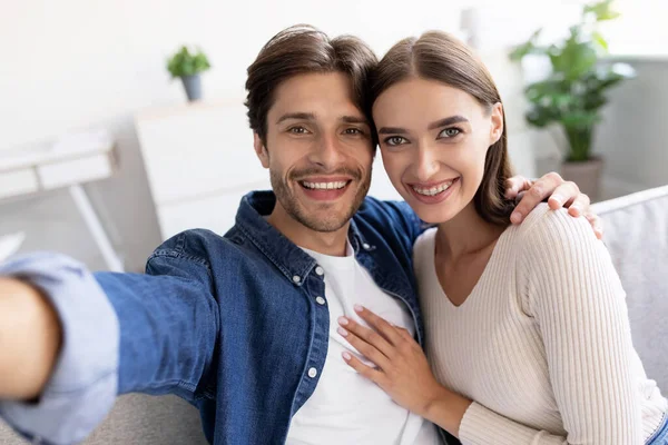 Sorrindo jovem europeia senhora e cara abraçando, olhar para webcam e tirar selfie no interior da sala de estar — Fotografia de Stock