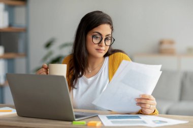 Merkez ofis. Belgeler ve laptopla çalışan konsantre Arap iş kadını elinde sıcak içecek bardağı tutuyor.