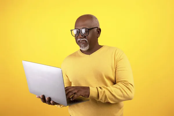 Senior zwarte man in bril met behulp van laptop computer, werken online op oranje studio achtergrond — Stockfoto
