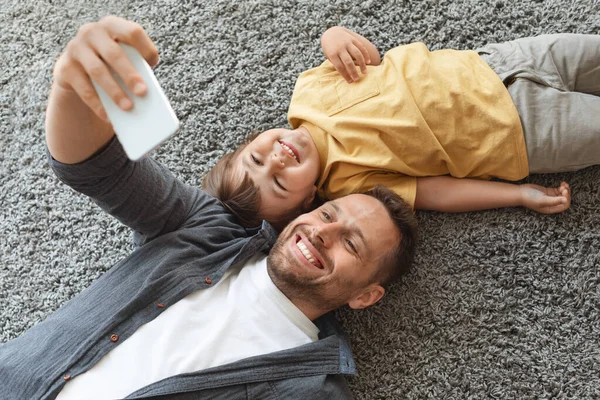 Feliz retrato de família. Jovem pai fazendo selfie com seu filho, sorrindo para smartphone, descansando juntos no chão — Fotografia de Stock