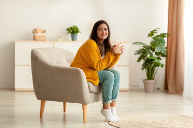 Pozitif Arap bir kadın elinde bir fincan sıcak kahveyle koltukta oturuyor ve kameraya gülümsüyor.