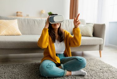 Modern VR gözlük takan neşeli Arap genç kadın oturma odasında oturmuş fotokopi alanını işaret ediyor.