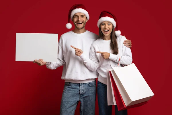 サンタ帽子をかぶったカップルがショッピングバッグを保持し、ブランクプラカードを指す — ストック写真