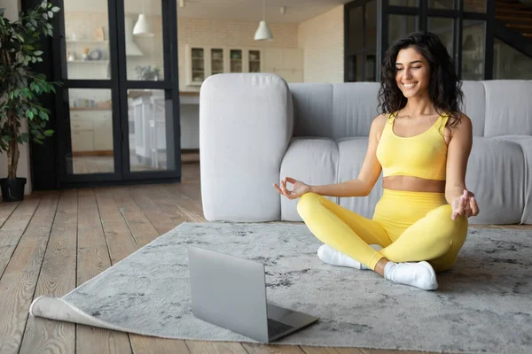 Спокійна тисячолітня жінка в спортивному одязі, сидячи в позі лотоса, має онлайн-клас йоги, роздумуючи біля ноутбука вдома — стокове фото