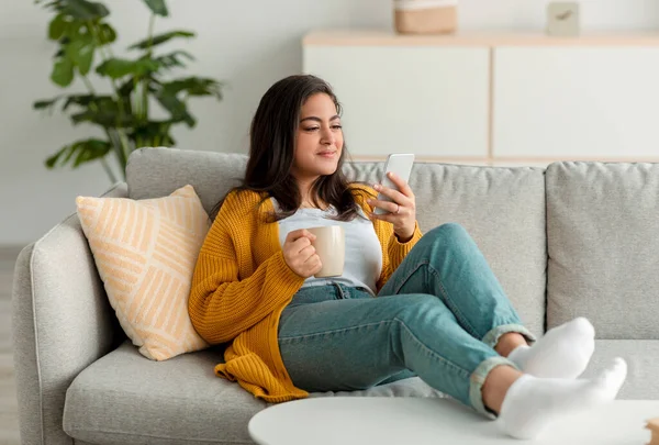 Livskoncept på nätet. Avslappnad arabiska dam med modern smartphone och njuta av kaffe hemma, sitter på soffan, fritt utrymme — Stockfoto