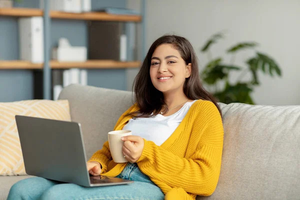 Film kijken of chatten op sociale netwerken. Positieve arabische vrouw met laptop en kopje thee of koffie rustend op de bank — Stockfoto