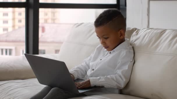 Preto preteen menino digitando no laptop enquanto sentado no sofá em casa — Vídeo de Stock