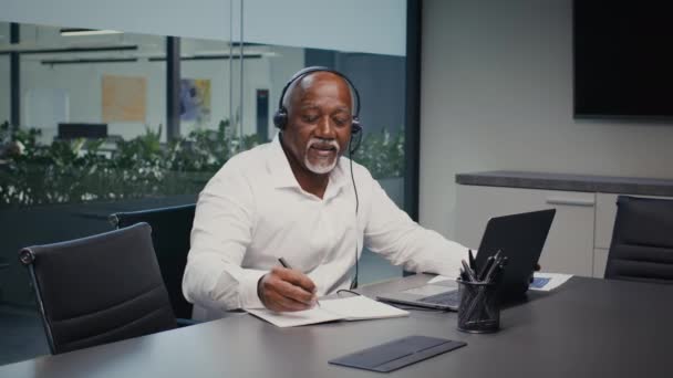 Черный бизнесмен в наушниках делает заметки во время работы с ноутбуком в офисе — стоковое видео