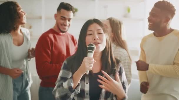 Fröhliche Party zu Hause. Junge positive asiatische Frau singt Lied mit Mikrofon, Gruppe fröhlich diverser Freundinnen tanzt — Stockvideo