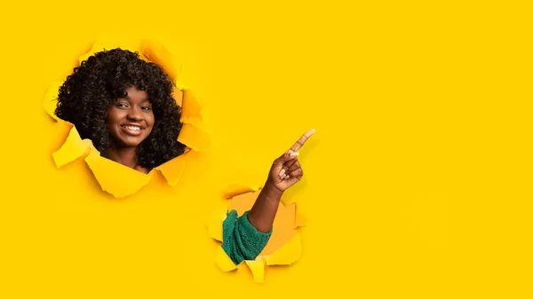 Mulher encaracolada afro-americana adulta feliz olha através do buraco no papel amarelo e aponta seu dedo para o espaço vazio — Fotografia de Stock