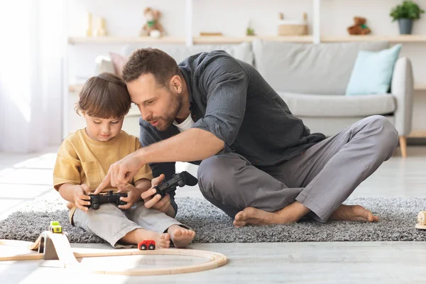 Νεαρός μπαμπάς να εξηγεί στον μικρό γιο πώς να παίζει με το χειριστήριο, να μαθαίνει στο παιδί του να παίζει βιντεοπαιχνίδια, να διασκεδάζει μαζί. — Φωτογραφία Αρχείου