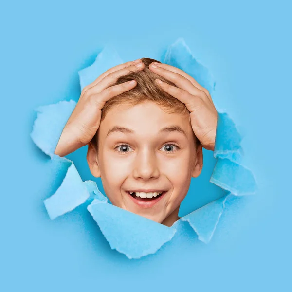 Shocked feliz emocionado adolescente europeo sosteniendo sus manos detrás de la cabeza mirando a través de agujero en papel azul —  Fotos de Stock