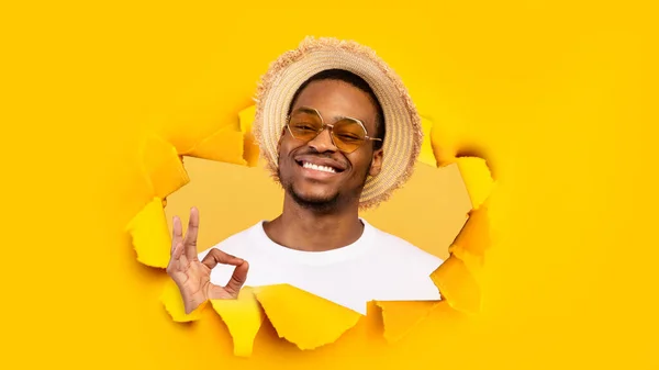 Веселий розслаблений тисячолітній чорний хлопець в сонцезахисних окулярах і капелюсі дивиться через отвір в жовтому папері і показує знак ОК — стокове фото