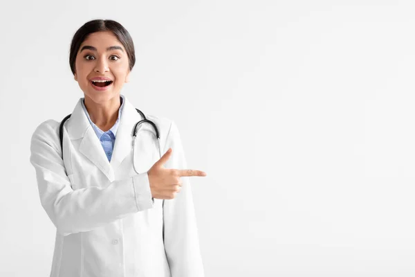 Surprised cheerful young indian woman in white coat with open mouth shows finger to empty space — Stock Photo, Image