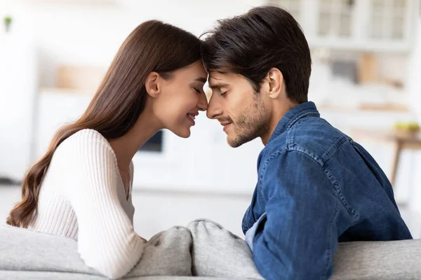 Feliz mujer caucásica milenaria apoyada frente a su marido sentado en el sofá y disfrutando de un momento romántico — Foto de Stock