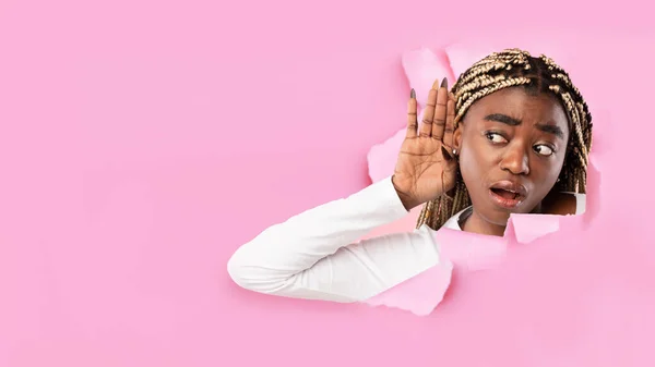 Surpreendido interessado millennial preto fêmea com boca aberta coloca a mão para orelha e escuta através de buraco em papel rosa — Fotografia de Stock