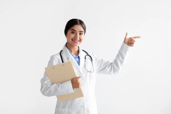 Glimlachende jonge indiaanse dame in witte jas met tablet toont vinger op lege ruimte, geïsoleerd op witte achtergrond — Stockfoto