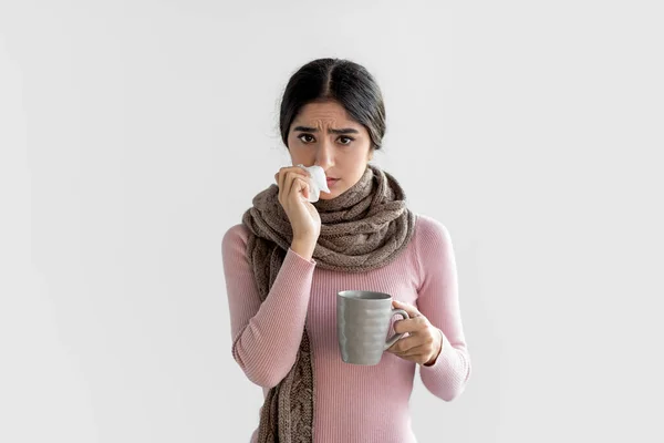 Сумна нещаслива індійська леді тисячоліття в шарфі з чашкою гарячого напою, витираючи її ніс серветкою — стокове фото