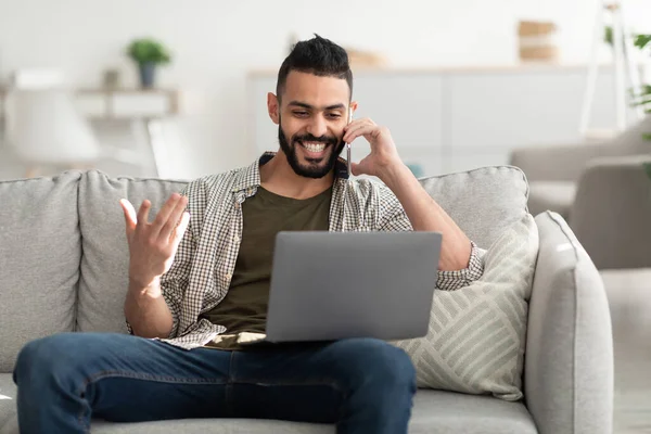 Hombre árabe joven positivo usando el ordenador portátil, teniendo conversación de teléfono móvil, trabajando en casa — Foto de Stock
