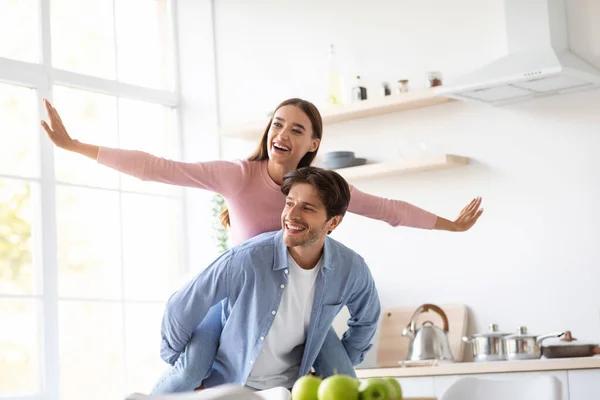 Feliz jovem marido carrega sua esposa nas costas, divirta-se juntos na internet da cozinha — Fotografia de Stock