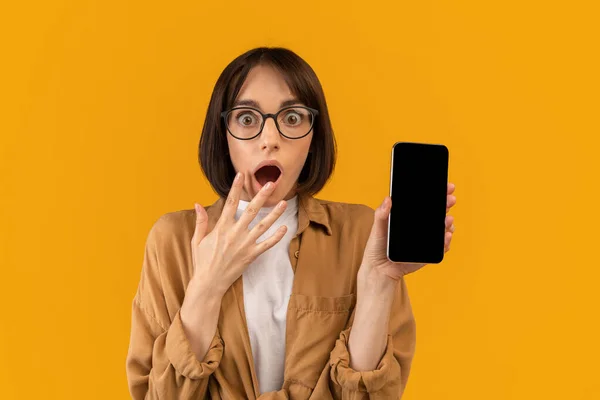 Emotionele dame toont smartphone met leeg scherm en het bedekken van haar mond in shock op gele achtergrond, mockup — Stockfoto