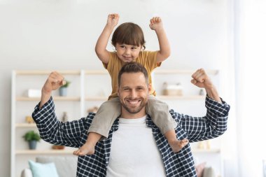 Aile gücü. Küçük oğlunu omuzlarında taşıyan genç ve güçlü bir adam, kaslarını gösteriyor ve kameraya gülümsüyor.