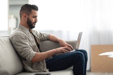 Çevrimiçi iş, iletişim. Genç bir Arap video görüşmesi yapıyor, dizüstü bilgisayarda internette geziniyor, evdeki koltukta oturuyor.