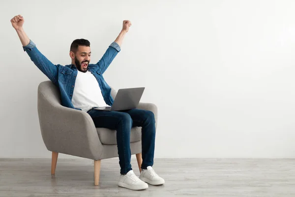 Emocionado joven árabe sentado en sillón con portátil, celebrando el éxito del trabajo, ganar la lotería en línea, espacio para copiar — Foto de Stock