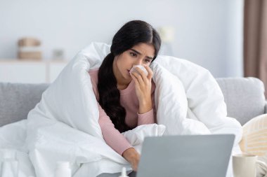 Mutsuz genç Hintli kadın beyaz battaniyeye sarılmış, soğuktan acı çekiyor, kanepeye oturuyor, aletlere bakıyor.