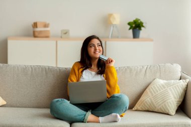 Alışverişkolik. Kredi kartı tutan ve dizüstü bilgisayar kullanan düşünceli Arap kadın internetten alım yapmayı düşünüyor.