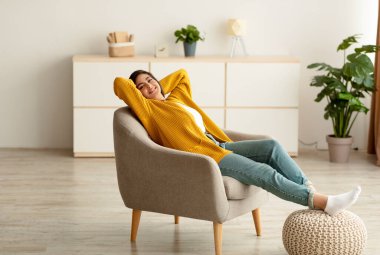 Mutlu Ortadoğulu kadın evinde rahat koltukta dinleniyor, Arap kadın elleri başının arkasında oturuyor.