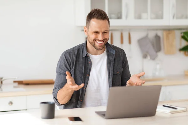 Reunión de negocios virtual. Alegre joven hombre de vídeo de chat con sus colegas en línea, hablando con el ordenador portátil webcam en la cocina — Foto de Stock