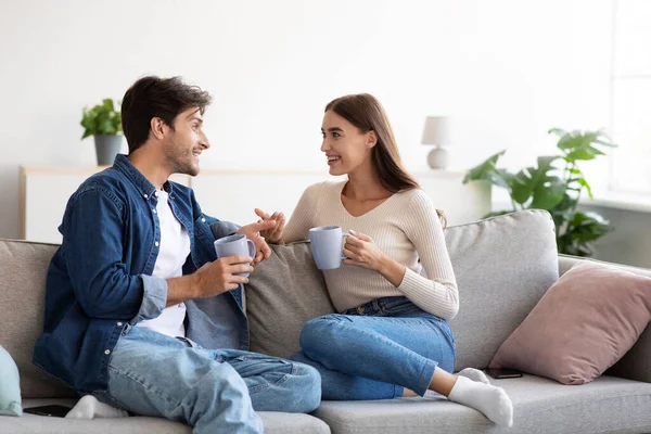Cheerful lovers young european guy and lady, with cups of hot drink talk in room interior — Stock Photo, Image