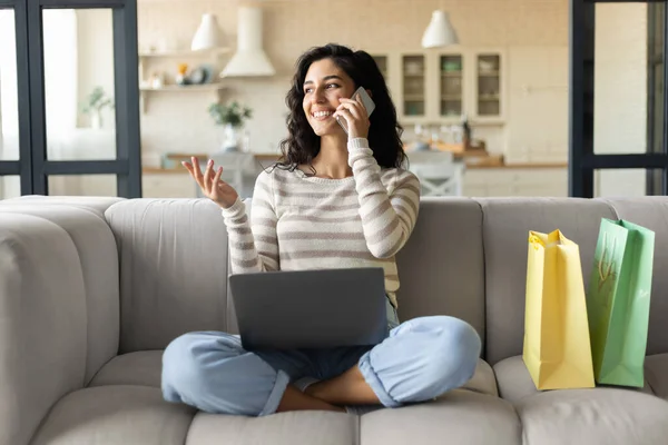 Preciosa joven morena con bolsas de regalo de compras en línea en el ordenador portátil, confirmando el pedido web en el teléfono inteligente desde casa — Foto de Stock